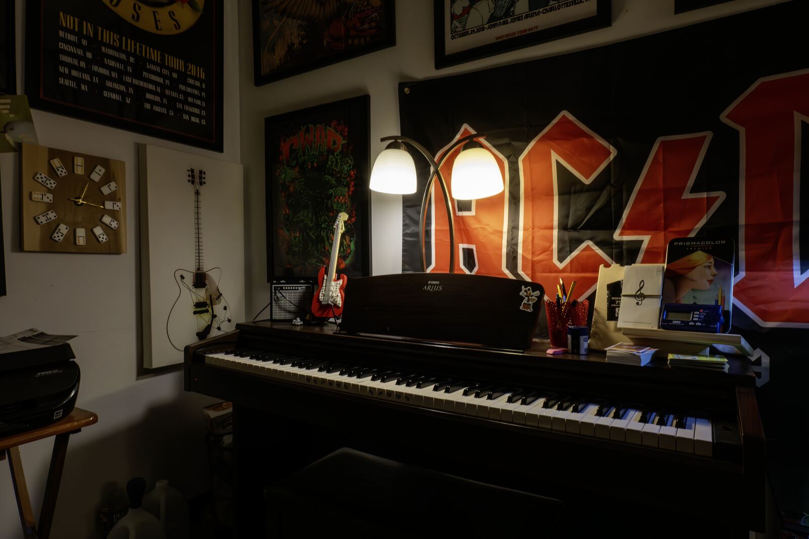 A digital piano lit by a twin-shade lamp, under a large AC/DC flag and framed posters